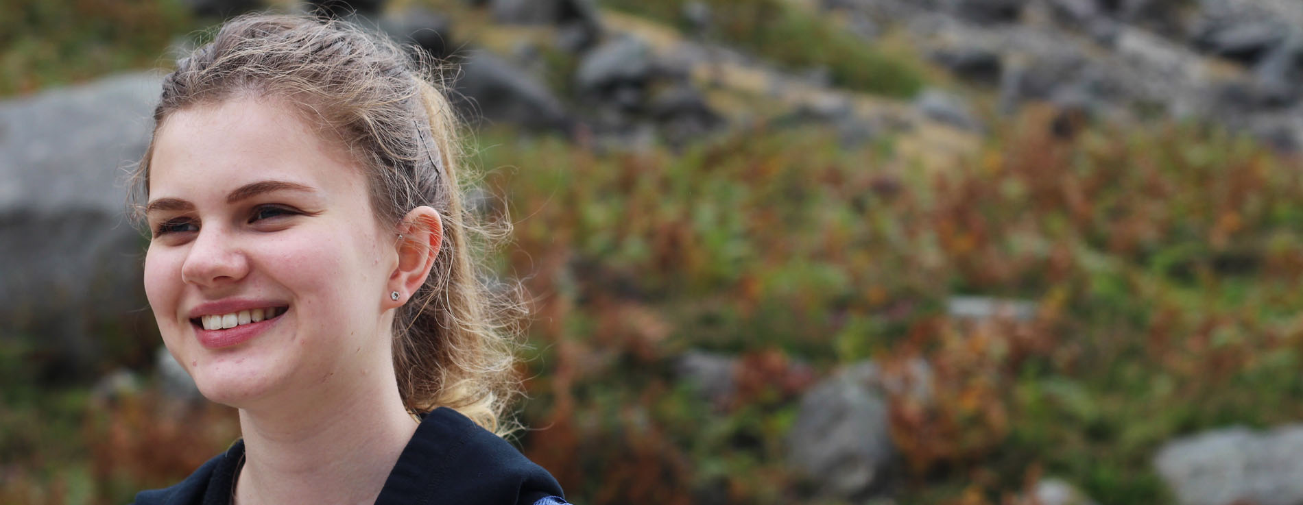 A young woman with curly blonde hair smiles outdoors, standing in front of a blurred background of rocks, green grass, and brown plants.