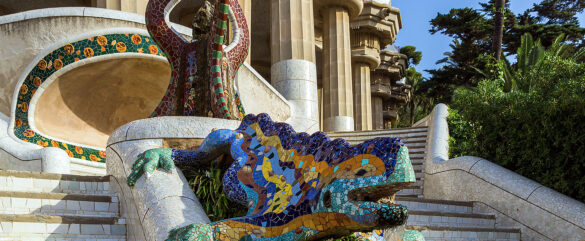 Colorful mosaic lizard sculpture beside curved stone steps at Park Güell in Barcelona, Spain, with lush greenery and unique architectural columns in the background.