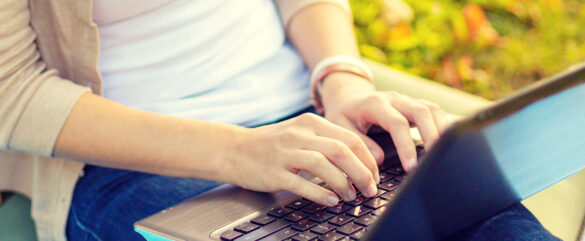 A person sitting outdoors types on a laptop placed on their lap. The person is wearing a light-colored shirt and blue jeans, and the background is blurred with green and yellow tones.