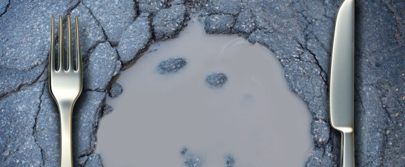 A fork and knife are placed on either side of a pothole filled with murky water on an asphalt road, resembling a dinner plate.
