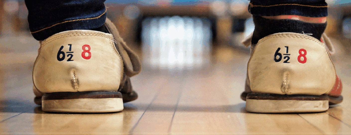 Close-up of two peoples feet in worn bowling shoes standing on a bowling lane, with bowling pins visible in the blurred background. The shoes display the size 6½ 8 on the heels.