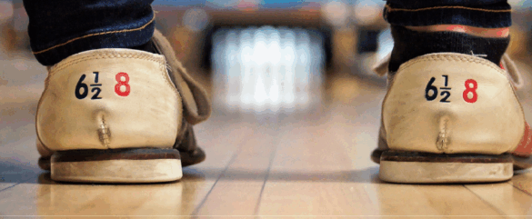 Close-up of two peoples feet in worn bowling shoes standing on a bowling lane, with bowling pins visible in the blurred background. The shoes display the size 6½ 8 on the heels.