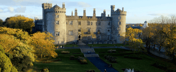 A large stone castle with round towers stands behind neatly manicured gardens and trees with autumn leaves, under a blue sky with scattered clouds. People walk along paths leading up to the castle entrance.